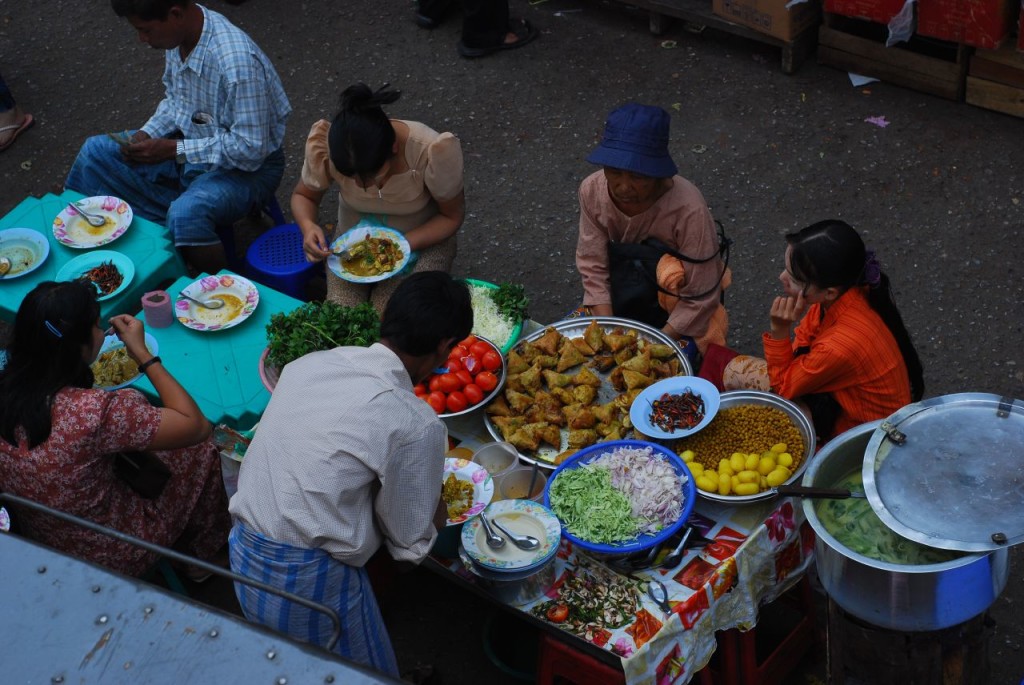 People eating outside on small benches at a table filled with large platters of colorful food.