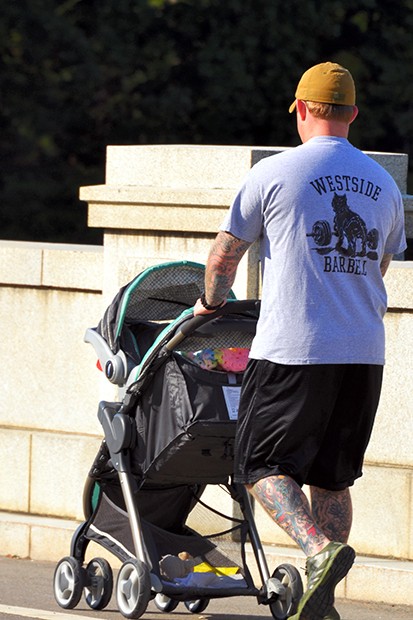 Man with tattooed legs and arms in athletic clothing, pushing a baby stroller.
