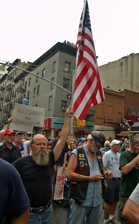 A bearded man holds high a large American flag in his left hand amidst a protest, a sign reads no clubhouse for jihadists.