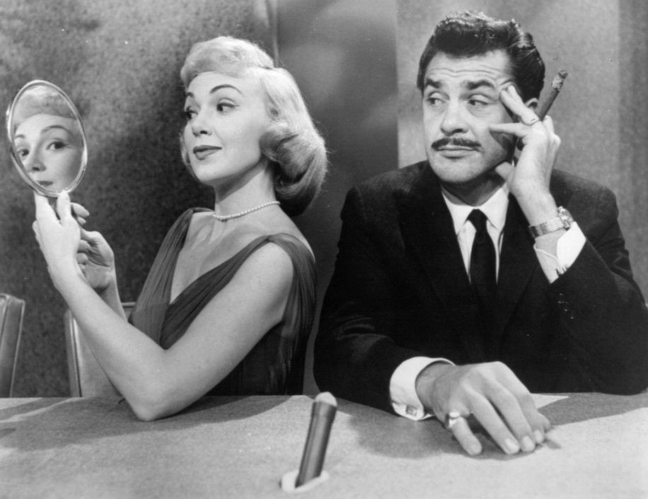 Two people sitting next to each other, one looking into a mirror and the other looking at the first while holding a cigar.