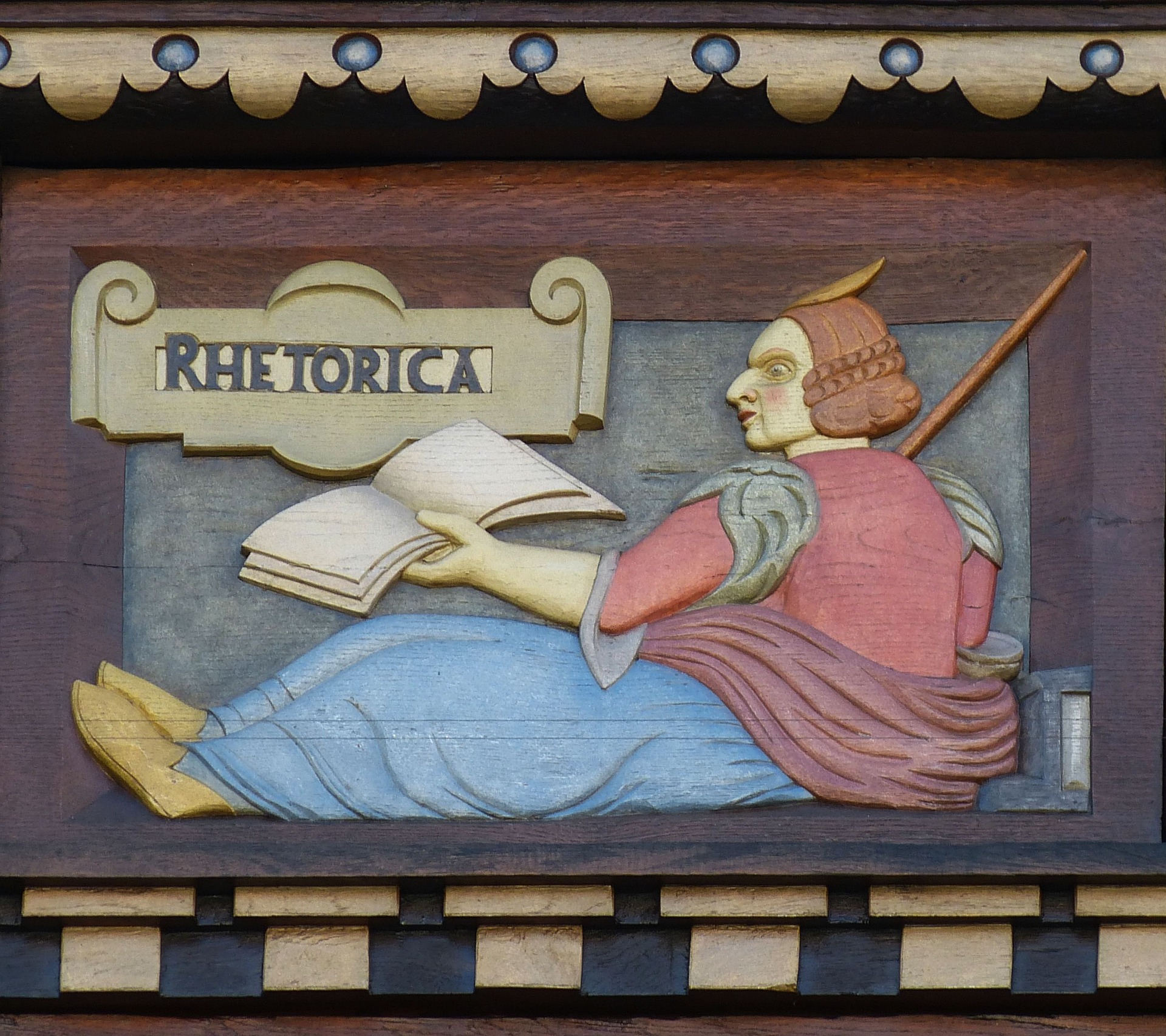 wood carving showing a seated figure holding a book, labeled "Rhetorica"