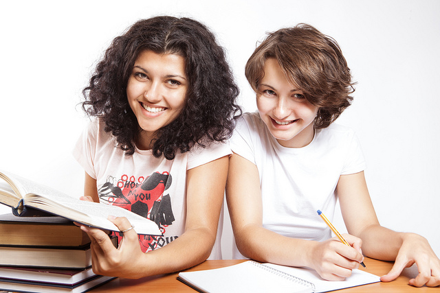 Photo of two young women seated at a table with books and papers in front of them, smiling into the camera