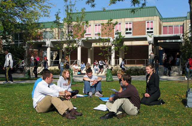 Photo of a group of students sitting in a circle on a lawn outside of a campus building, while other students stream around them