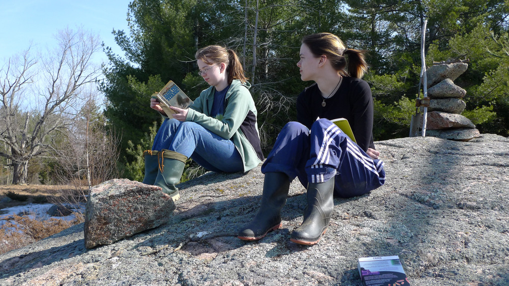 Photo of two young women sitting on a rock. One is reading a book.