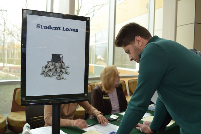 A male student leans over a table with a sign on it that reads, "Student Loans." Two women are seated behind the table.