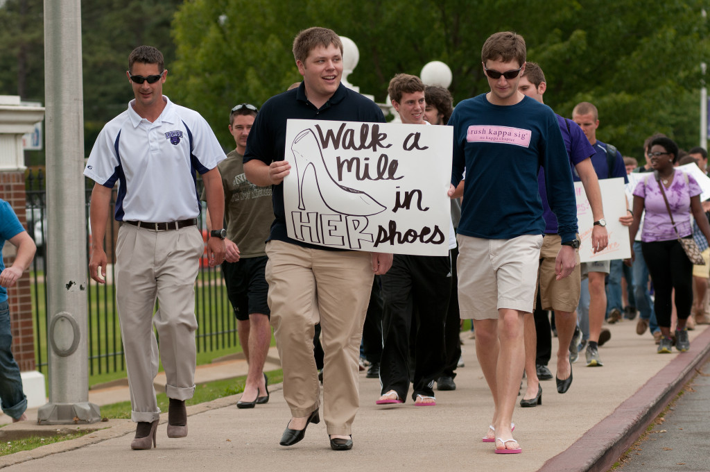 A group of male students is walking; most are wearing women's shoes, and one holds a sign that reads, "Walk a Mile in Her Shoes."
