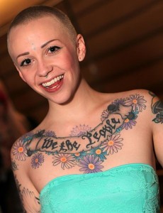 Photo of woman with a closely cropped haircut and facial piercings. She wears a strapless top to reveal a large tattoo across her chest reading "We the People" surrounded by flowers.