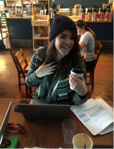 Photo of a girl seated in a coffee shop, smiling, holding a coffee cup and her other hand is resting against her chest.