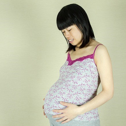 Very pregnant Asian woman in a casual dress peering downward with both hands resting on her very large belly.