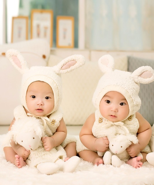 Foto de bebés gemelos muy lindos de ascendencia asiática sentados uno al lado del otro con ropa de bebé encapuchada rematada con orejas de conejo.