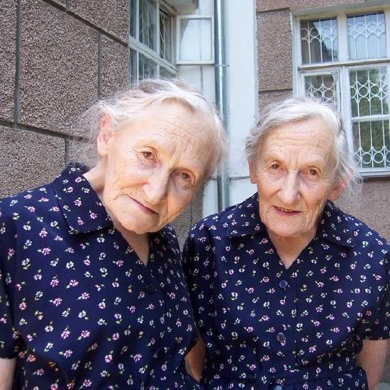 Photo of a pair of elderly identical twin women in identical dresses with identical faces, and gray white hair, facing camera.