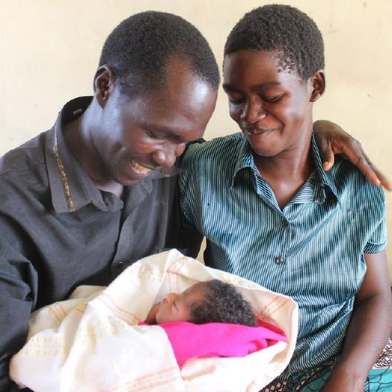 Well dressed, African couple in Western clothing, smiling at infant held in man's arm, his other arm around woman's shoulder.