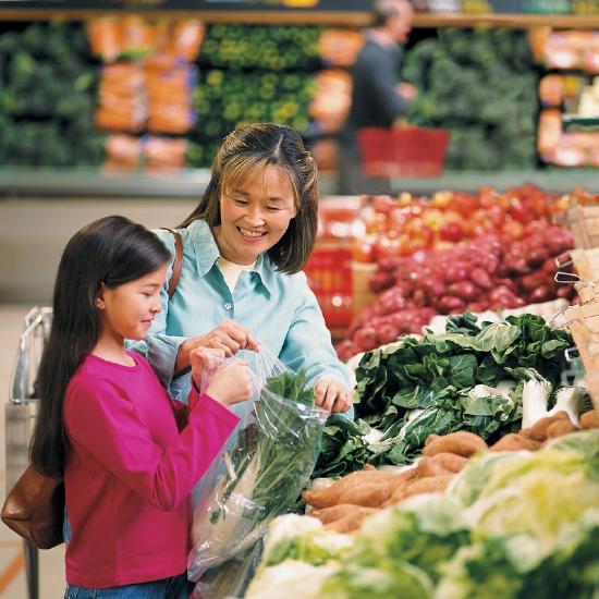 Asian mother in produce isle of a Western supermarket smiling at her young daughter as they put leafy bok choy in a bag.