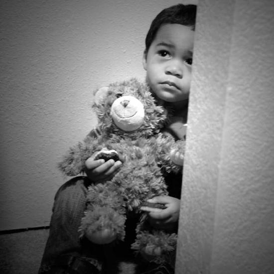 Black youngster about five or six years old sitting in a corner holding stuffed animal with worried look on his handsome face.
