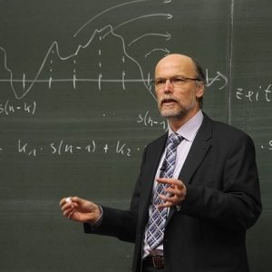 Una imagen estereotipada de un profesor -un hombre blanco, de mediana edad, con gafas y barba, vestido con abrigo y corbata se levanta con tiza en la mano frente a una pizarra que muestra una fórmula matemática.