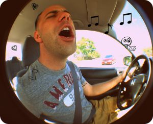 A young man sits behind the wheel of a car with his eyes closed as he sings along with the radio.