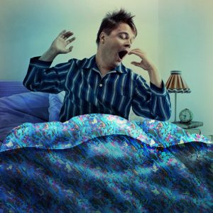A man dressed in pajamas sits up in bed as he stretches and yawns.