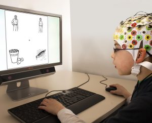 Un niño con gorra EEG usa un mouse y un teclado mientras mira imágenes en un monitor de computadora durante un experimento.