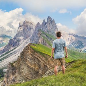 A man stands in an alpine meadow and looks into the distance at the high mountain peaks." title="A man stands in an alpine meadow and looks into the distance at the high mountain peaks.