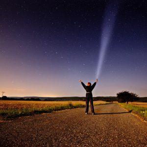 A woman stands in the middle of a country road at night and reaches towards the star-filled sky at night.