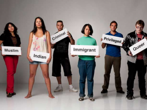 A group of people stand holding signs labeling them as others perceive them. The signs include, 'Muslim', 'Indian', 'Criminal', 'Immigrant', 'Privileged', and 'Queer'.