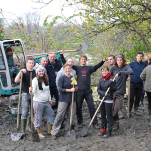 Un grupo de hombres y mujeres se unen en un campo fangoso con palas y carretillas mientras participan en un proyecto de voluntariado al aire libre.