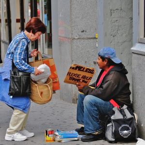 A woman stops on the sidewalk to offer food to a man holding a sign reading 'Homeless, please help Thank you.'