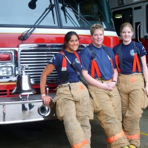 Tres mujeres integrantes de un equipo de bomberos y rescate se paran frente a un motor de bomberos con ropa de seguridad contra incendios.