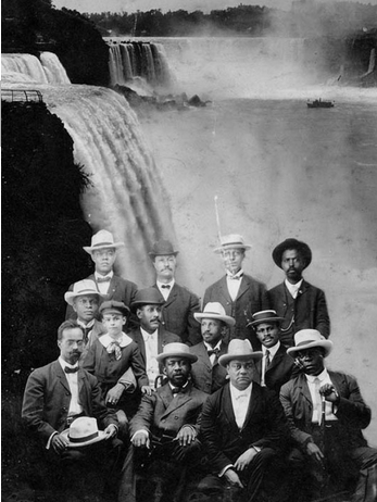 Photograph of the members at a Niagara movement meeting in Fort Erie, Canada, 1905. Photograph is taken in a studio with the image of the Niagara falls added into the background.