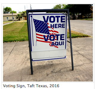 2016 photograph of a metal sign outside a polling place in Taft, Texas. The sign contains a graphic of the American flag and the phrase "Vote here" in both English and Spanish.