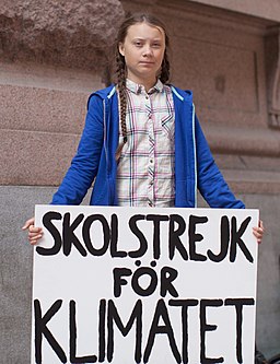 Photograph of Greta Thunberg outside the Swedish parliament building, August 2018. She is holding a sign that reads "Skolstrejk för klimatet," meaning, "school strike for climate."