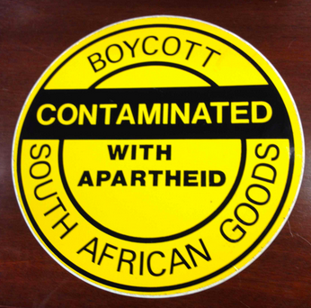 Circular yellow sign on a wood background, with black text on the sign reading "Boycott South African Goods: Contaminated with Apartheid."