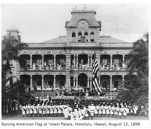 The U.S. flag is raised over Iolani Palace, Honolulu, Hawaii in a formal event on August 12, 1898. The space in front of the palace is filled with U.S. marines in dress uniforms.