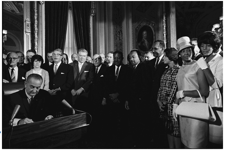 Black-and-white photograph of President Lyndon B. Johnson signing the Voting Rights Act of 1965, while Martin Luther King, Jr. and other dignitaries and activists look on. Photograph taken by Yoichi Okamoto.