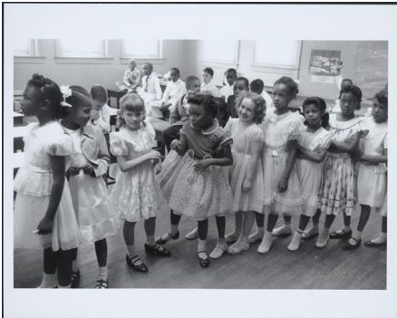 1955 black-and-white photograph by Thomas J. O'Halloran, showing a racially integrated classroom in Barnard School, Washington, D.C. A number of elementary-age girls, both Black and white, are clustered in the foreground, and a group of Black and white boys the same age sit at their desks in the background.