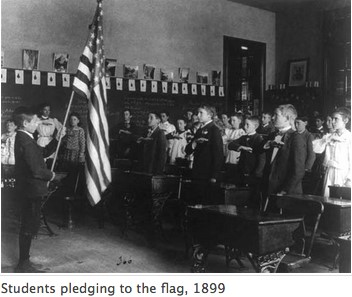 Fotografía en blanco y negro de 1899 que muestra a los alumnos de un aula de primaria frente a la bandera estadounidense y recitando el Juramento de Lealtades con las manos sobre el corazón. Fotografía tomada por Frances Benjamin Johnston.