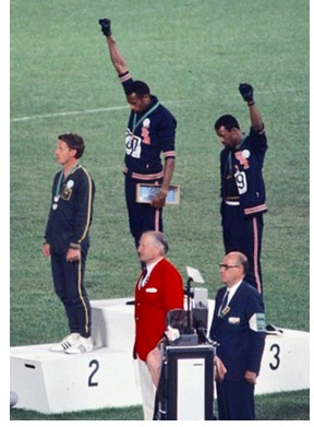 Photograph of the award podium for the men's 200-meter sprint at the 1968 Mexico City Olympic Games. Australian Peter Norman is in second place, and Black Americans Tommie Smith and John Carlos, in first and third place, respectively, are barefoot on the podium, bowing their heads, and each raising one fist in a black glove while their anthem is played.