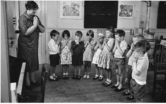 Fotografía en blanco y negro de 1963 de una clase de alumnos blancos de primaria siendo conducidos a recitar la Oración del Señor por su maestro.