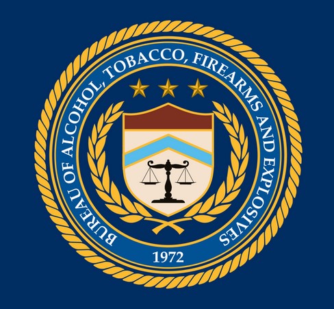 Graphic of the U.S. Bureau of Alcohol, Tobacco, Firearms, and Explosives, showing a medallion with the name of the agency surrounding a shield that pictures two balanced scales, surrounded by a gold laurel wreath and 3 gold stars.
