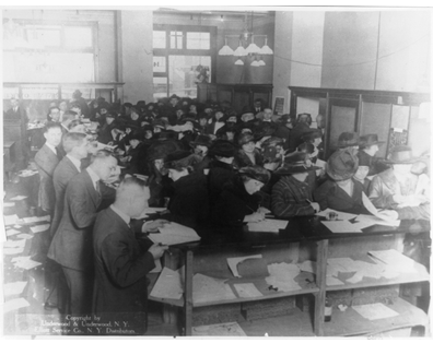 Black-and-white photograph by Underwood & Underwood, taken circa 1920, showing a number of men and women crowded at long desks in an Internal Revenue Service office to fill out tax forms.