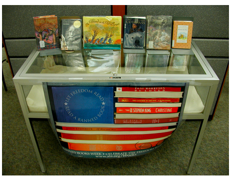Glass display holding several frequently-challenged books, at Carmichael Library, Montevallo, Alabama. Physical copies of Harry Potter and the Sorcerer's Stone, The Giver, The Wind in the Willows, The Golden Compass, The Wizard of Oz, and Winnie-the-Pooh are displayed on top of the case. A banner hangs from the case, designed to resemble the U.S. flag with the message "Let freedom read: read a banned book" written in white on the blue unison, the white stripes represented by the sides of books, and the red stripes represented by the spines of books such as Beloved, The Catcher in the Rye, Christine, To Kill a Mockingbird, and Of Mice and Men.