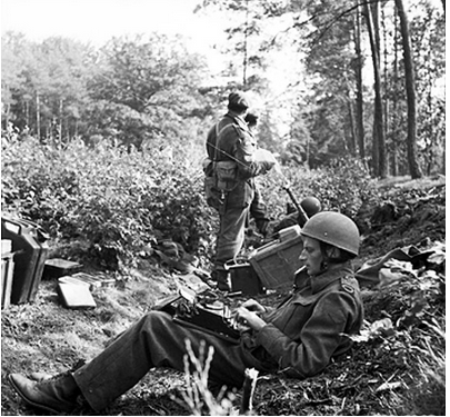 Black-and-white photograph of war correspondent Alan Wood typing his dispatch in a wood outside Arnhem, the Netherlands, on September 18, 1944. Three members of the 1st British Airborne Division are in frame behind Wood.