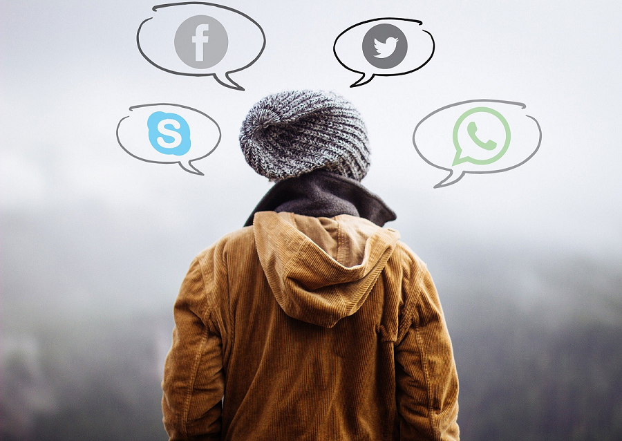 Photograph of a person wearing a winter coat and hat, facing away from the camera. Four speech bubbles, each holding the logo of a different social media company (Snapchat, Twitter, Facebook, Whatsapp), radiate out from the person's head.