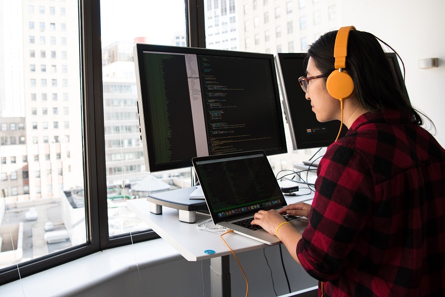 A woman wearing headphones is working on a coding project, using both a laptop and a desktop computer.
