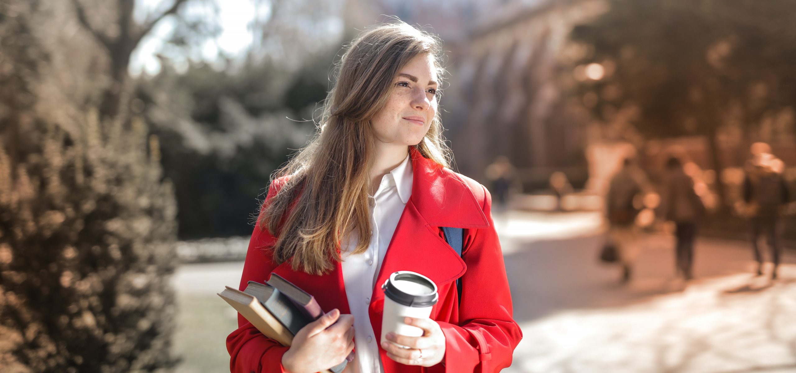 Mujer vestida con abrigo rojo, sosteniendo libro y café mientras camina por terrenos universitarios