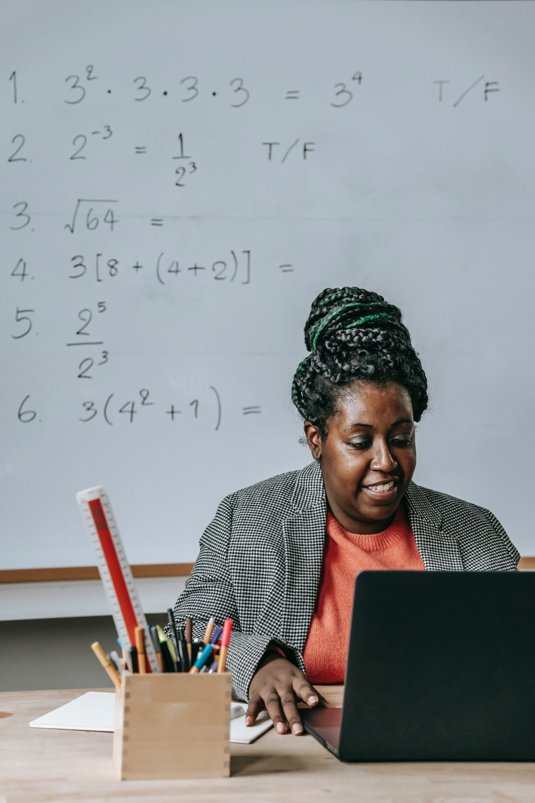 Mujer negra sentada en el escritorio con laoptop con pizarra blanca de ecuaciones detrás de ella