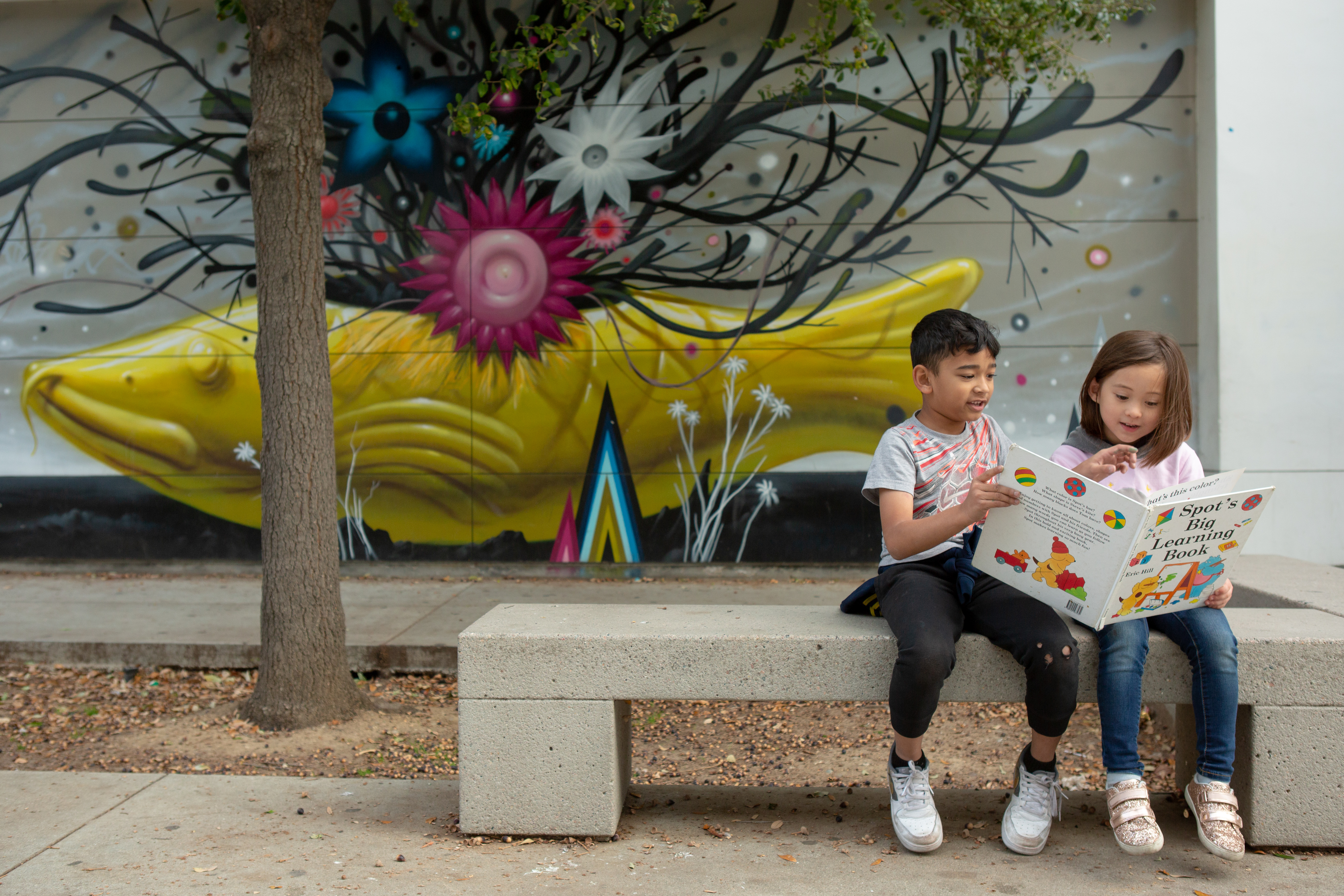 Two children sit on a bench in front of a mural holding a book together. 