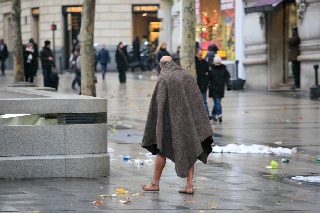 Homeless man wearing a blanket with barefeet walking on a cold street, with others walking around in heavy coats.
