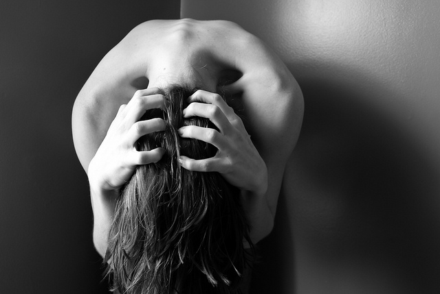 Black and white photograph of a woman who is hunched over with her hair covering her face and grabbing the back of her hands with her head, depicting anguish.