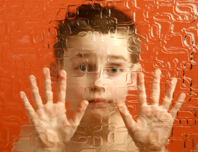 Photograph of an autistic boy looking through a fuzzy glass window with his hands pressed against the glass.